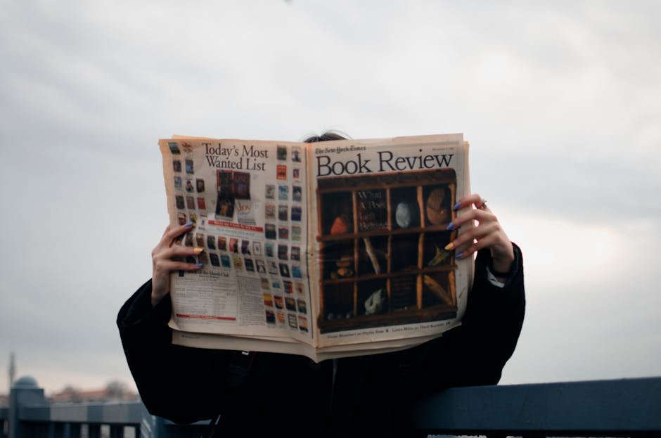 Individual holding a book review newspaper with colorful nails against a cloudy sky backdrop.