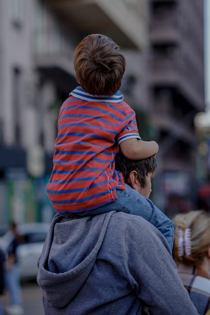 A father carries his child on his shoulders while walking down a busy urban street.