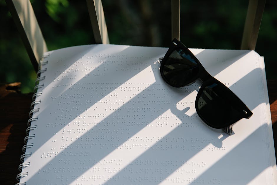 Spiral-bound Braille book with sunglasses casting striped shadows, outdoors in natural light.