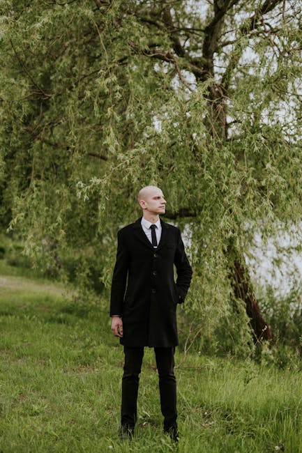Adult man in a black suit and tie standing in a lush green park.