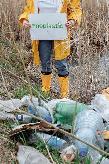 Person in yellow raincoat holding #NoPlastic sign near littered area advocating environmental care.