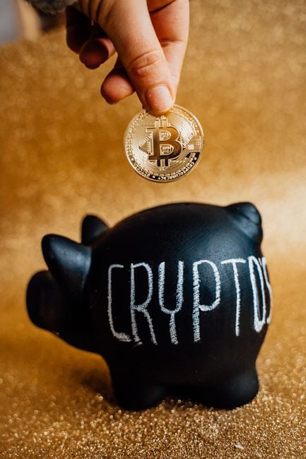 A hand places a Bitcoin into a piggy bank labeled 'Crypto', symbolizing savings and investment.