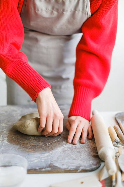 Unrecognizable female in apron working with clay on wooden board while standing at table with rolling pin in professional workshop