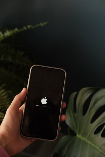 Person holding a smartphone displaying an Apple logo indoors with a leafy plant in the background.
