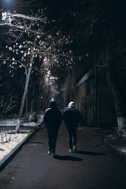 Two friends walk through a snowy, dimly lit street in Almaty at night, embracing winter's natural beauty.