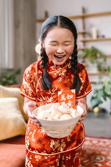 A young girl in a red traditional outfit joyfully holding a bowl of fortune cookies indoors.