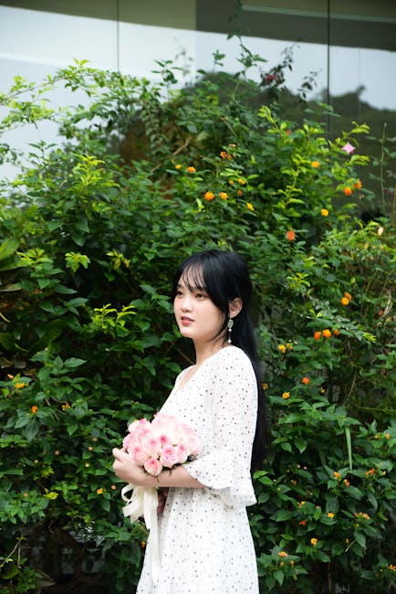 Woman with pink roses stands amidst lush greenery outdoors.