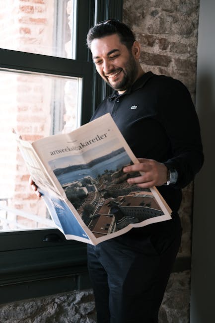 Adult man with beard reading a newspaper by a window in Istanbul, smiling.