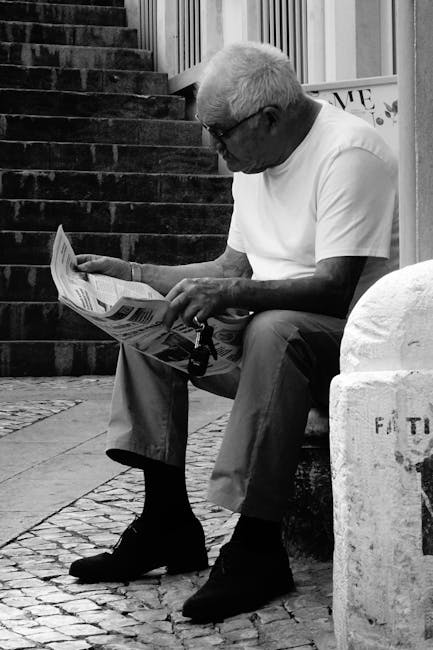A senior man in sunglasses sits on a bench reading a newspaper beside stone steps.