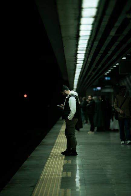 Young man using smartphone on a dark subway platform with overhead lights.