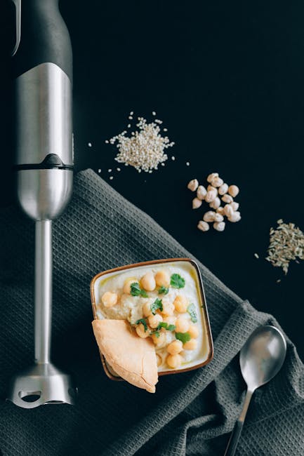 Flatlay of creamy hummus with chickpeas and pita, surrounded by blender and herbs.