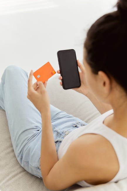 A woman sitting on a couch, making an online purchase using a credit card and smartphone.