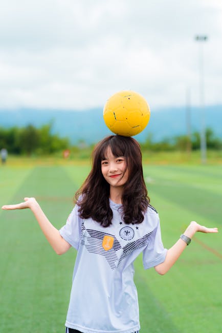 Young woman smiling while balancing a soccer ball on her head in a field.