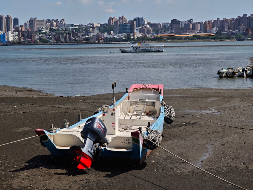 A fishing boat anchored on a muddy shore with a city skyline in the background, under a clear sky.