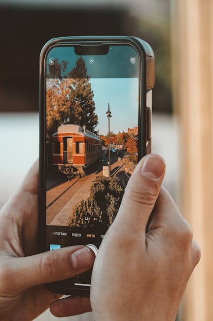 Hands holding a smartphone capturing a vintage train outdoors on a sunny day.