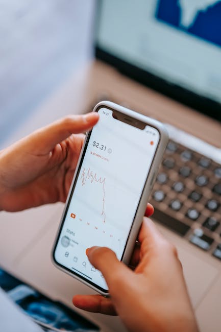 High angle of crop anonymous dealer using financial exchange application on cellphone with chart on screen against laptop