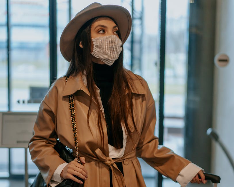 A fashionable woman in a brown coat and hat wearing a face mask inside an airport lobby.