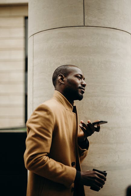 Side view of a stylish black man in a trench coat talking on his smartphone outdoors.