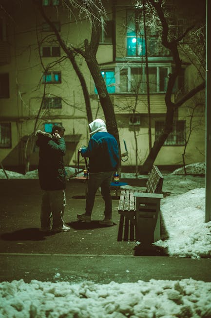 Two friends in winter clothing talking outside in a snowy urban setting at night.