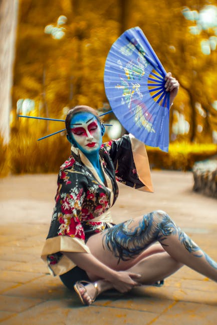 A performer in a colorful kimono with striking makeup poses outdoors with a fan.