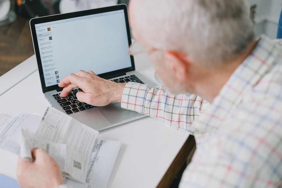 Elderly man at desk handling finances online with documents and laptop.
