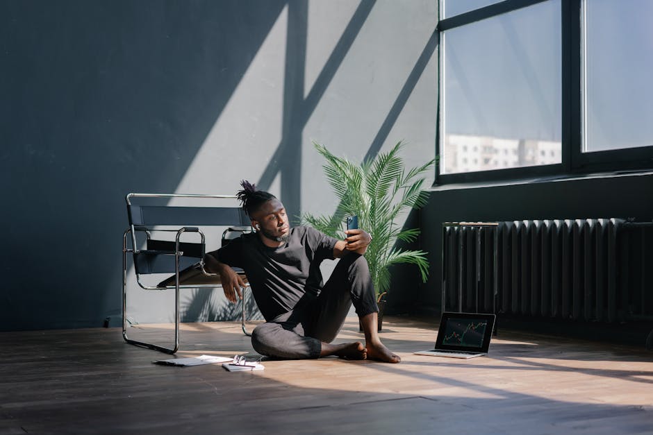 Man sitting on the floor, checking smartphone, with laptop showing stock market trends nearby.