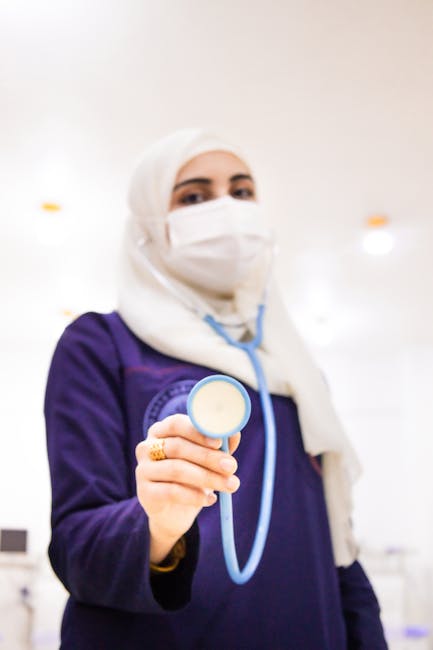 A female doctor in a hijab and mask holding a stethoscope in a medical setting.