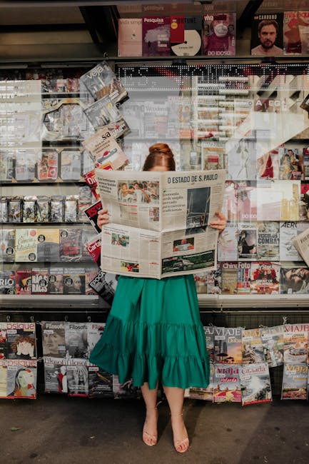 Woman in green dress reading newspaper at street magazine stand outdoors.