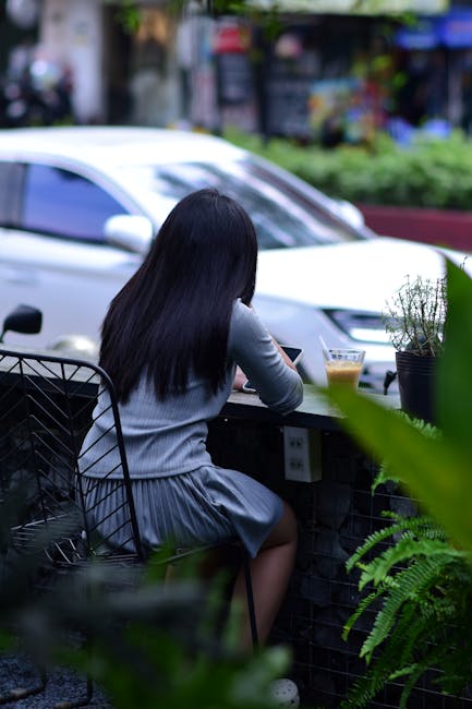 A woman sits outdoors at an urban cafe with a drink, surrounded by greenery and city life.