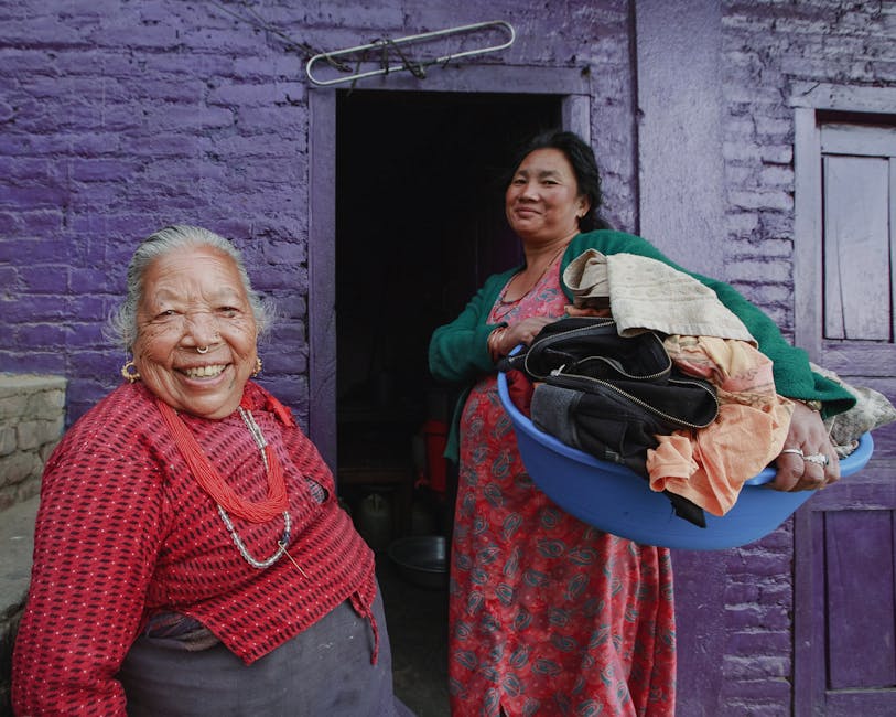Two women smiling outside a vibrant purple house, one holding a laundry basket.