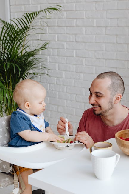A father joyfully feeds his baby in a high chair inside a cozy home environment.