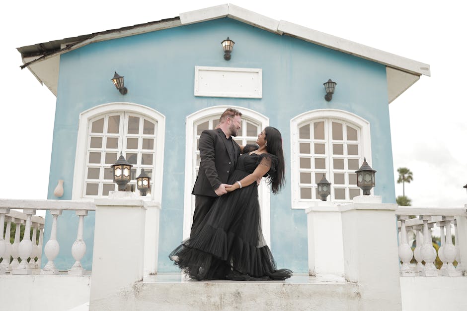 A couple in elegant black attire shares a romantic moment in front of a blue house.
