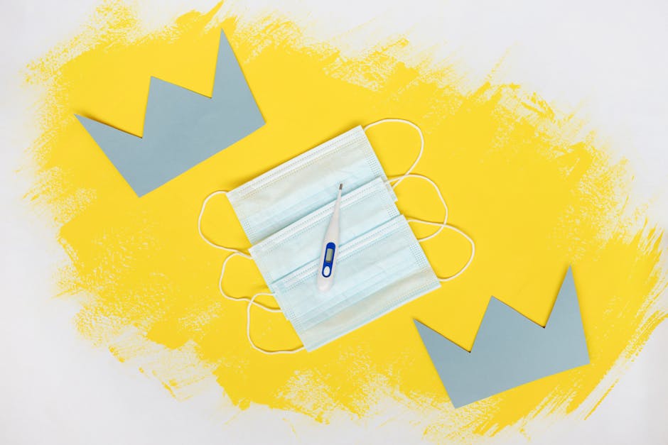 Artistic arrangement of a face mask and thermometer on yellow background.