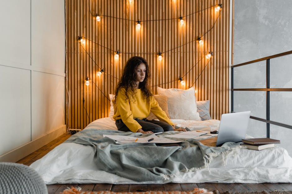 A woman working on a laptop in a cozy bedroom with string lights, creating a relaxed atmosphere.