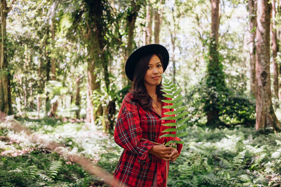 A fashionable woman in a plaid dress and hat stands amidst lush greenery holding a fern.
