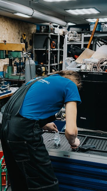 A technician repairs equipment in a cluttered workshop setting. Industrial tools surround them.