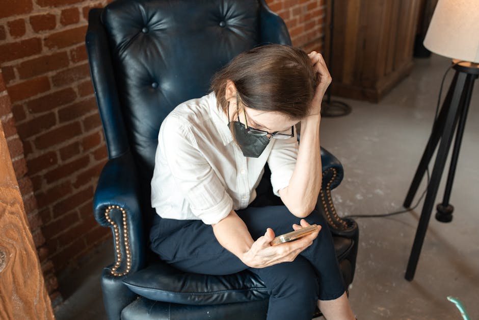 Woman wearing a mask, using a smartphone while seated in a blue armchair indoors.