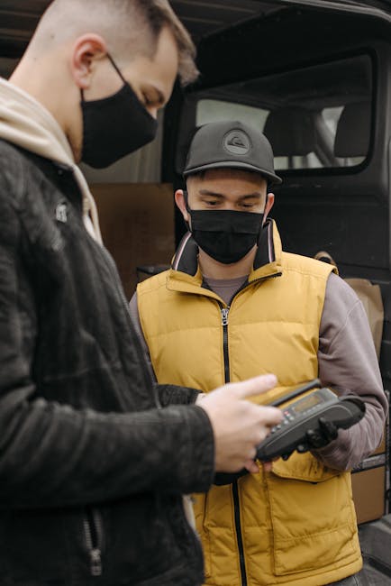 Masked deliveryman uses payment terminal for contactless service outside in urban setting.