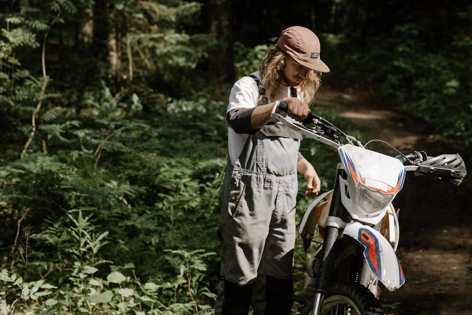 A man in overalls adjusts a dirt bike in a lush forest, highlighting outdoor adventure.