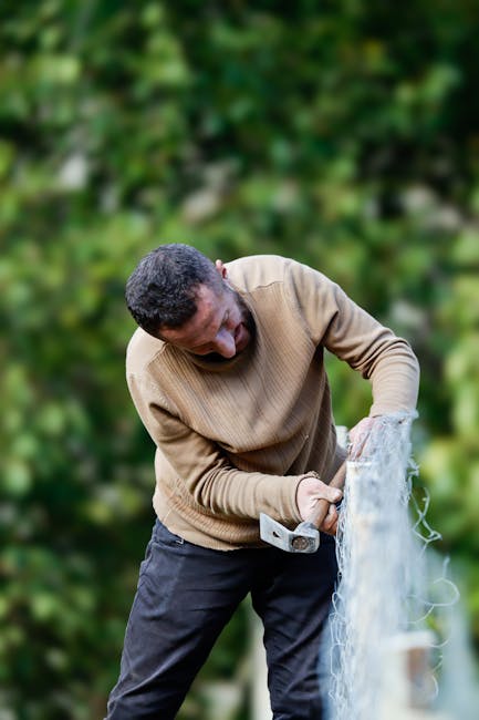 A man fixes a wire fence outdoors, surrounded by lush greenery.