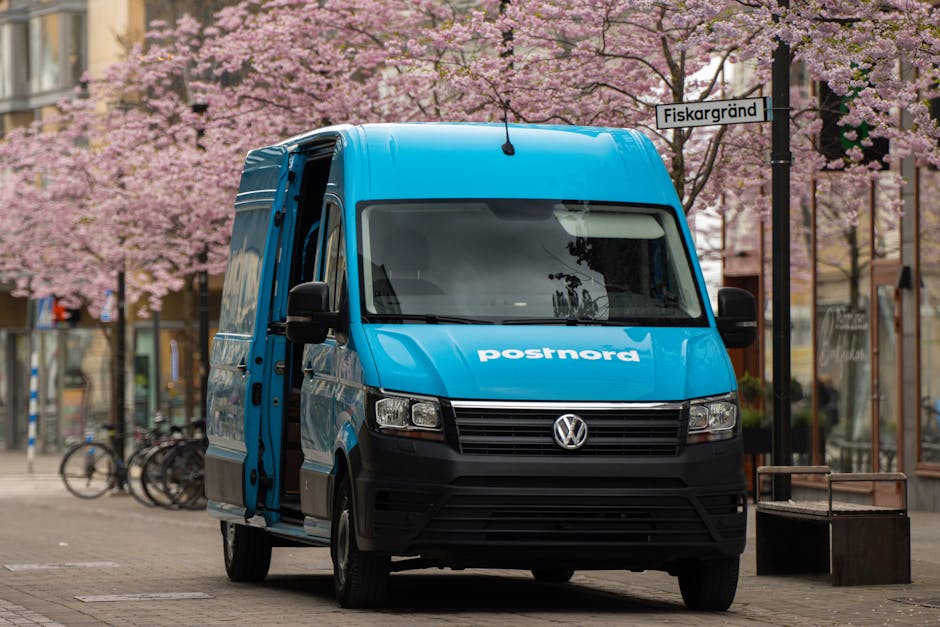 Blue PostNord van parked on a street in Jönköping, Sweden, lined with cherry blossoms.