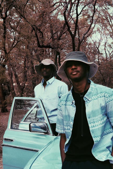 Two fashionable men wearing bucket hats stand by a vintage car in a wooded area.