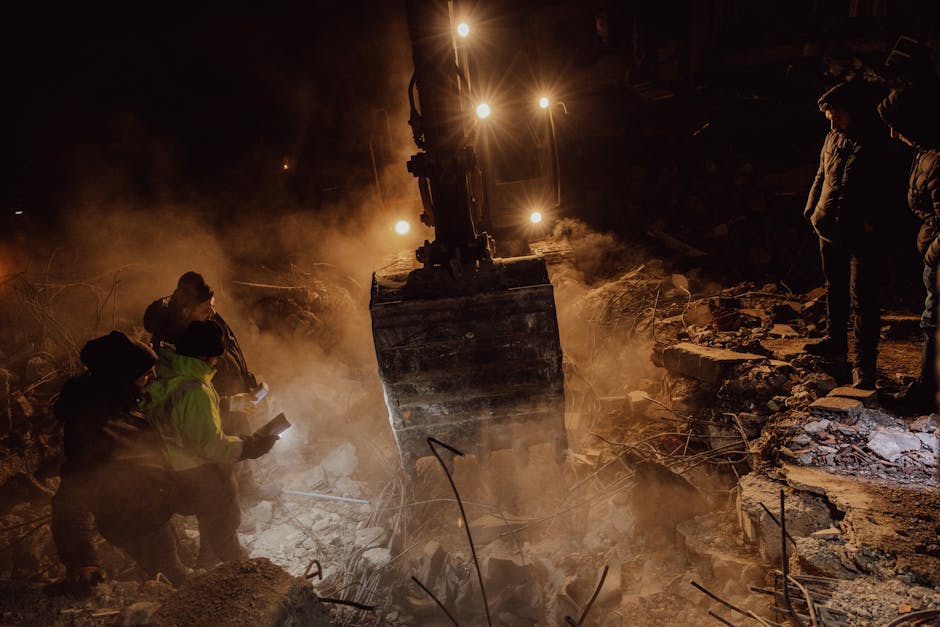 Emergency workers conduct a night rescue in debris after a disaster, illuminated by harsh lights.