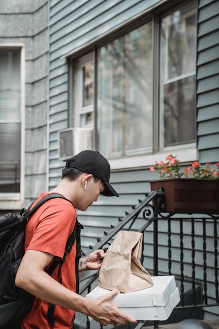 A young male delivery man carrying a pizza box and a paper bag, walking up residential steps.