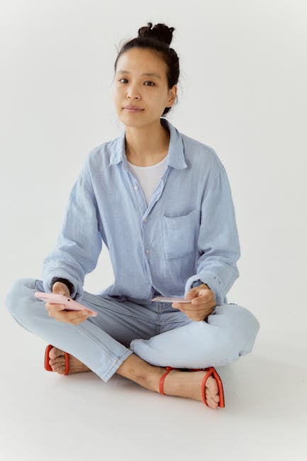 Young woman in casual blue attire sitting cross-legged holding a smartphone, on a white background.