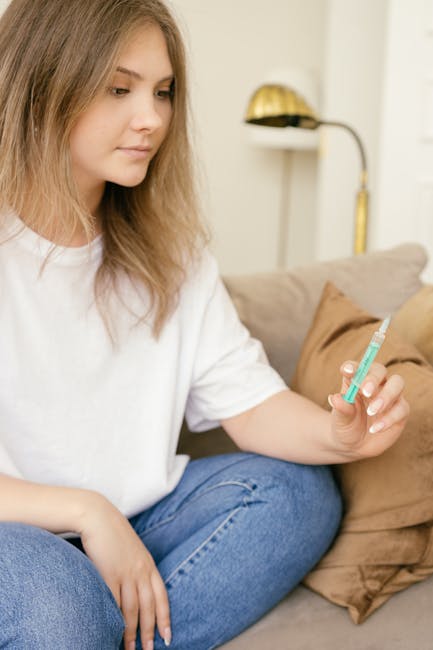 A young woman sitting on a sofa indoors, holding a syringe with a thoughtful expression.