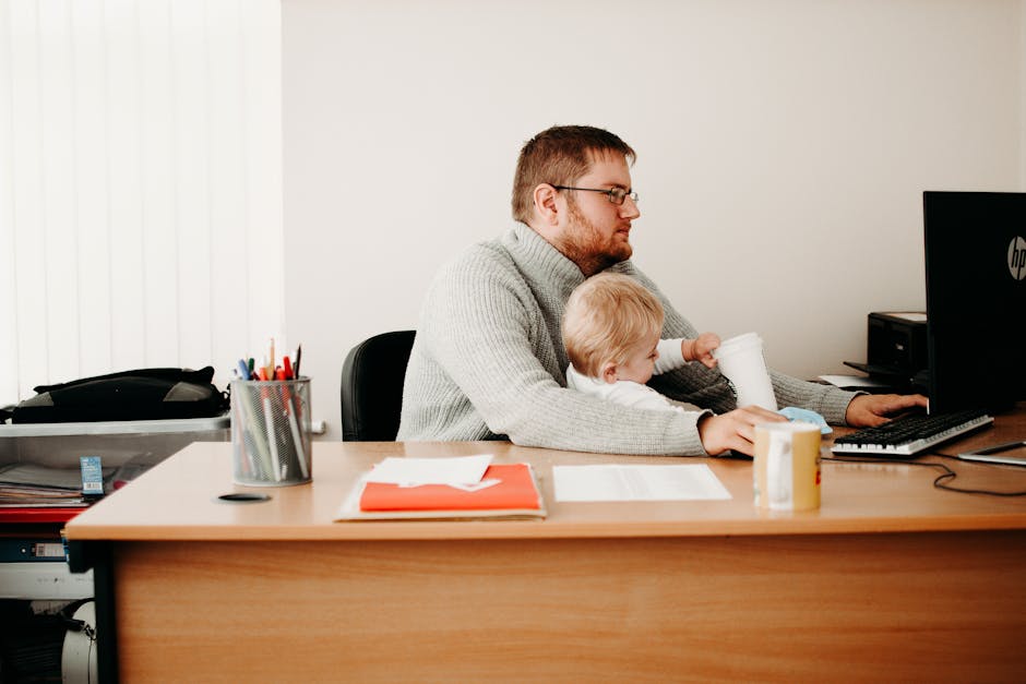 A father multitasks at his desk while holding his child, illustrating remote work and parenting.