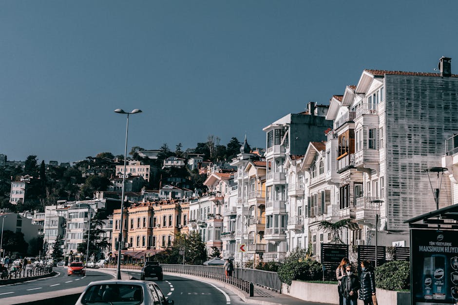 Asphalt driveway with cars riding along residential houses located on roadside against blue sky on street with people on bus stop in city