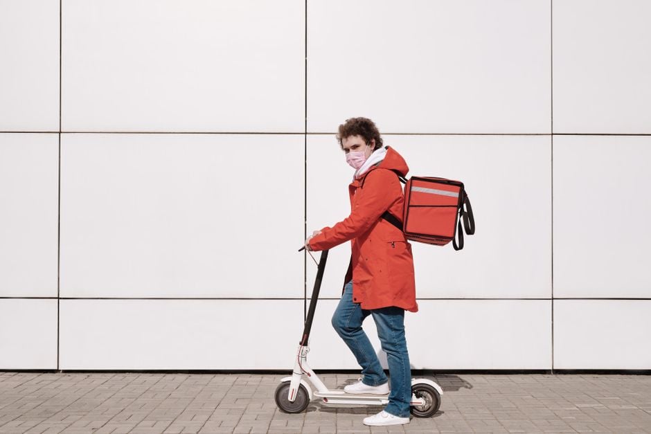 Man in red coat delivering food on scooter with safety mask outdoors.