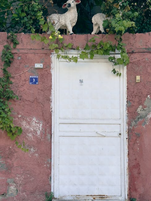 A rustic pink wall with a white door adorned by green vines and sheep sculptures in Halfeti, Türkiye.